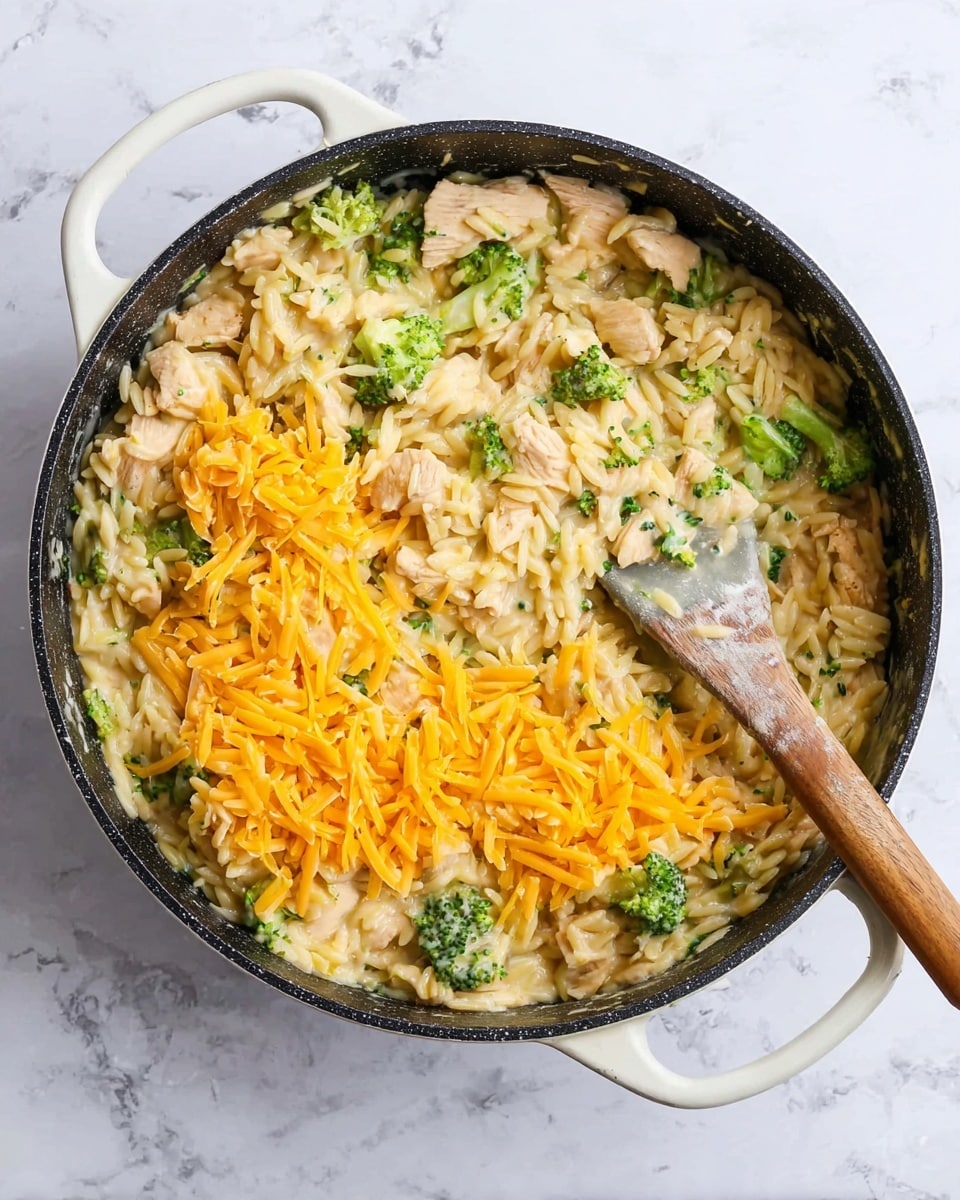 A white handled black pan filled with a creamy orzo pasta dish. The dish contains small pieces of light brown cooked chicken mixed with green broccoli florets throughout the pale creamy sauce. On top, there are several small piles of bright orange shredded cheddar cheese. A gray and wooden spatula is placed inside the pan, mixing the cheese into the pasta. The pan is sitting on a white marbled surface. photo taken with an iphone --ar 4:5 --v 7