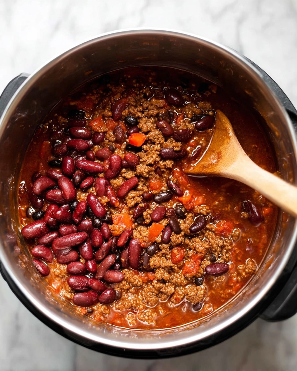 A close-up view inside a silver metal pot with black edges shows a mix of dark red kidney beans, black beans, and cooked ground meat in a rich, thick reddish-brown sauce with small orange carrot pieces. The beans and sauce cover most of the pot's inside. A light wooden spoon is partially plunged into the mixture on the right side of the pot, resting against the edge. The pot sits on a white marbled surface. photo taken with an iphone --ar 4:5 --v 7