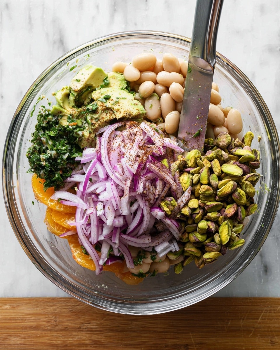 A clear glass bowl shows a colorful mix of ingredients layered inside; at the bottom, bright orange slices and avocado pieces add a fresh base, followed by a thick layer of white beans that fill most of the bowl, topped with a small pile of chopped green herbs on one side and a mound of thinly sliced purple onions dusted with a touch of dark spice on top. To the right, a pile of green pistachio nuts contrasts with the smooth beans and onions. A silver knife rests inside the bowl, and the whole scene is on a white marbled surface. Photo taken with an iphone --ar 4:5 --v 7