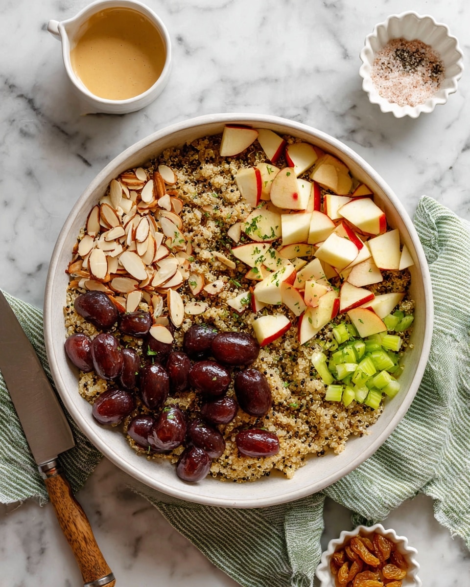 A large white bowl holds a layered salad with a base of cooked quinoa, light beige with black specks. On top, there are sections of thinly sliced almonds on the left, dark red grapes halved with shiny skin beside them, small chopped apple pieces with red skin and white flesh in the middle, and bright green chopped celery on the right. Golden raisins are scattered over the celery and apples. Fresh green chopped herbs are sprinkled on top, adding a touch of freshness. Around the bowl on the white marbled surface are a small white sauce cup with creamy tan dressing and a small white scalloped dish with mixed seasonings, likely pink salt and cracked black pepper. A knife with a white handle rests on a green and beige striped cloth under the bowl. Photo taken with an iphone --ar 4:5 --v 7